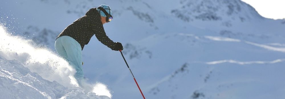 ACL-Injuries-in-Women Skier in helmet and goggles carving through fresh powder on a snowy slope with mountains in the background.