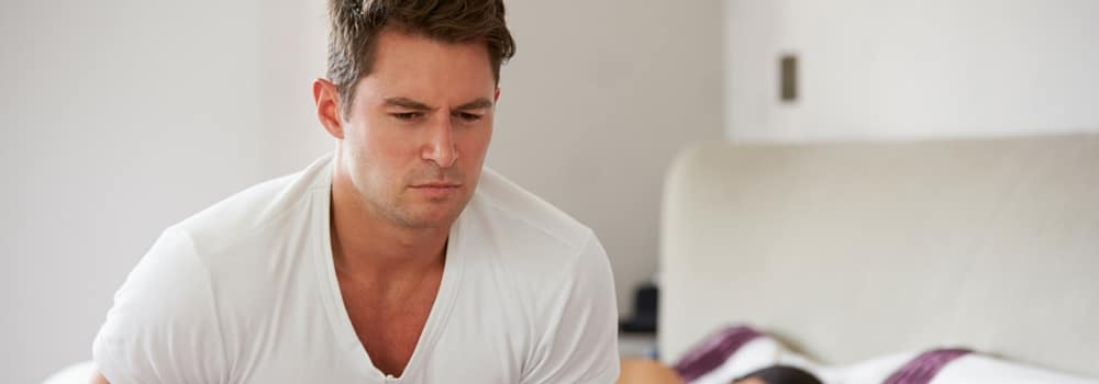 Mobile-IV-Nutrient-Therapy-case Man in a white t‑shirt sits on a bed, looking down with a concerned expression while a person lies under a blanket in the background.