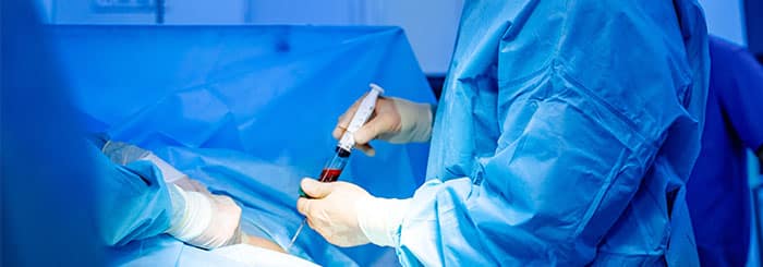 Operating room scene with clinicians in blue gowns using a syringe during a medical procedure.