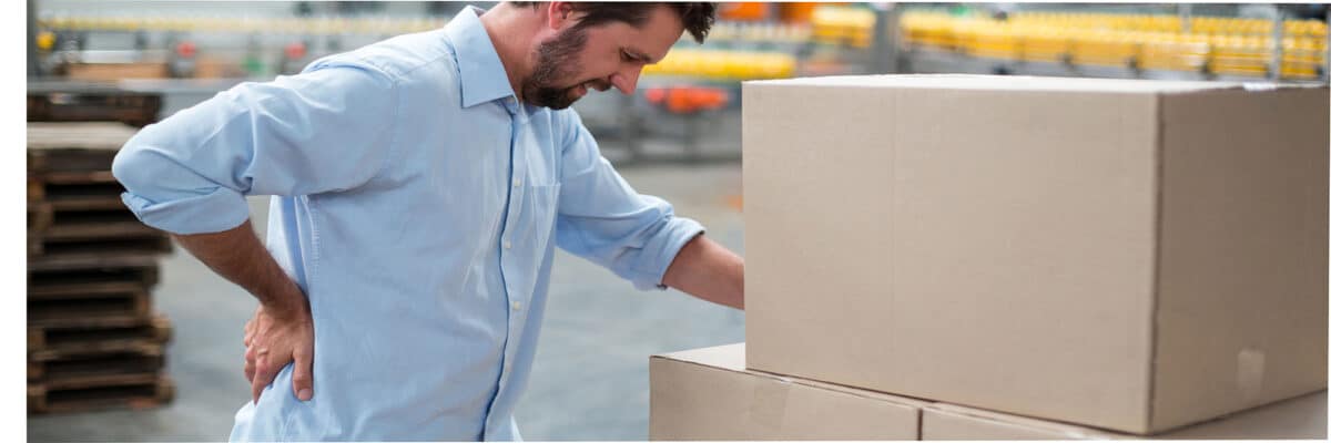 back-pic-1200×400 Man in a blue shirt bends toward stacked cardboard boxes in a warehouse, holding his lower back as if in pain or strain.