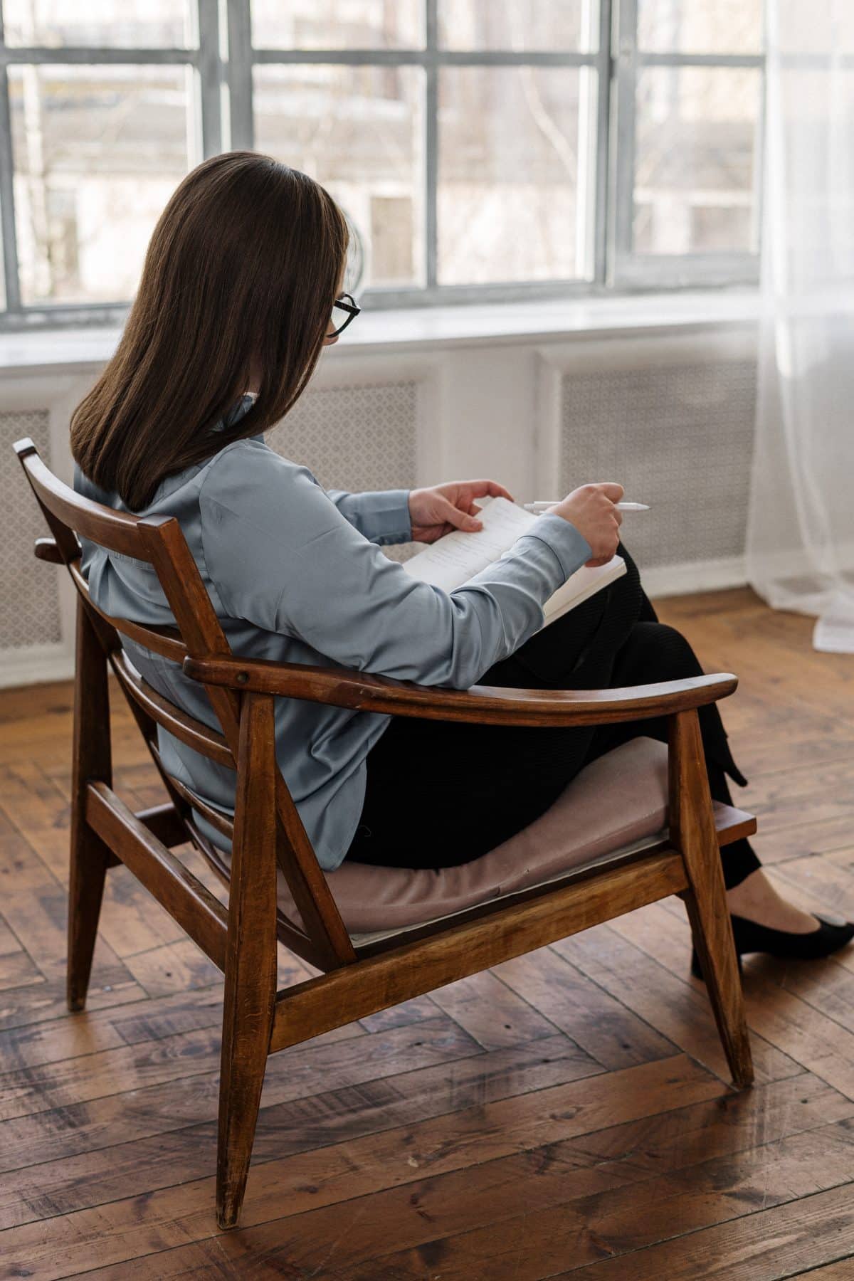 pexels-cottonbro-4101147-1200×1800 Person with brown hair and glasses sits in a wooden chair, writing in a notebook by a large window.
