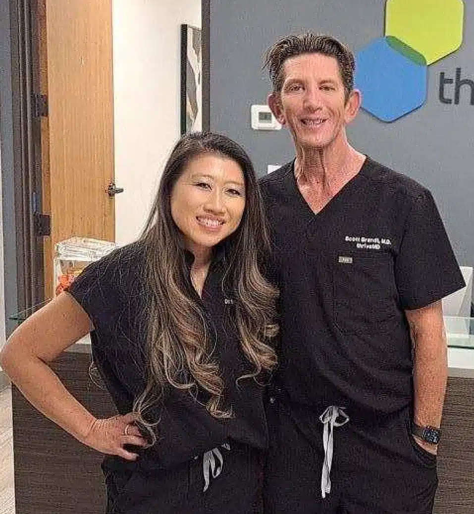 Portrait of a smiling medical professional in black scrubs standing in a clinical setting with medical equipment in the background.