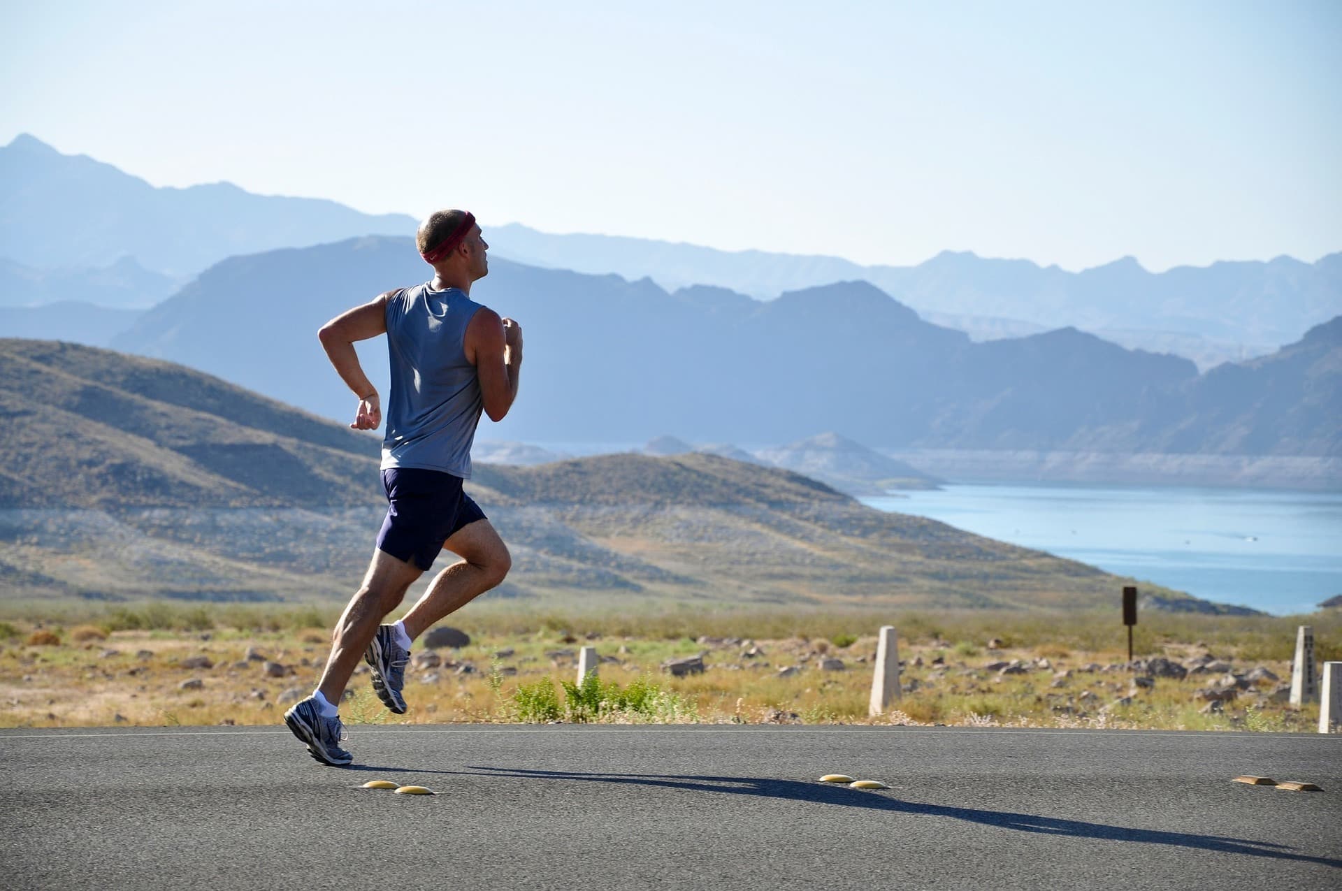 Male runner in a blue sleeveless shirt and shorts jogging on a paved road with a range of arid mountains in the background.